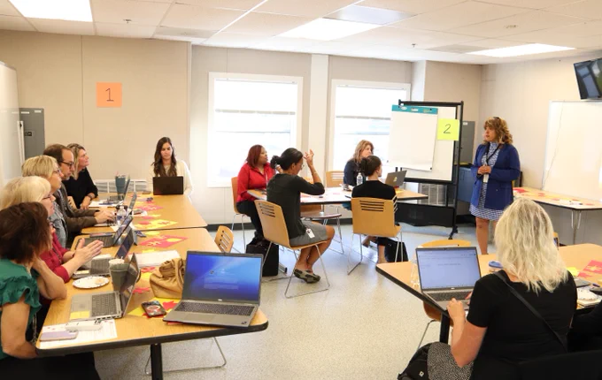 Photo of PLC attendees sitting at tables in a classroom with laptop computers in front of them. They are listening to a speaker at the front of the room.