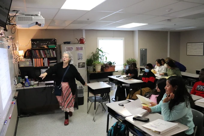 Photo of a classroom. Students are sitting at desks listening to a teacher at the front of the room who is pointing at the whiteboard.