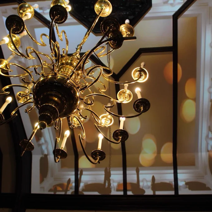 A golden chandelier holding over 13 artificial candles seen from below, the lights are reflecting on the white and wooden beamed ceiling of the room.