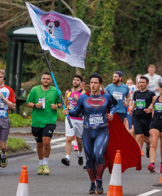 A photo of a participant at the 2024 Bath Half. He is wearing a Superman fancy dress costume and carrying a flag for Cots for Tots.