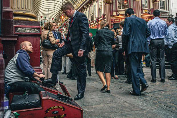A person gets their shoes shined at a London City outdoor shoe shine station, surrounded by bustling pedestrians in a covered alleyway.