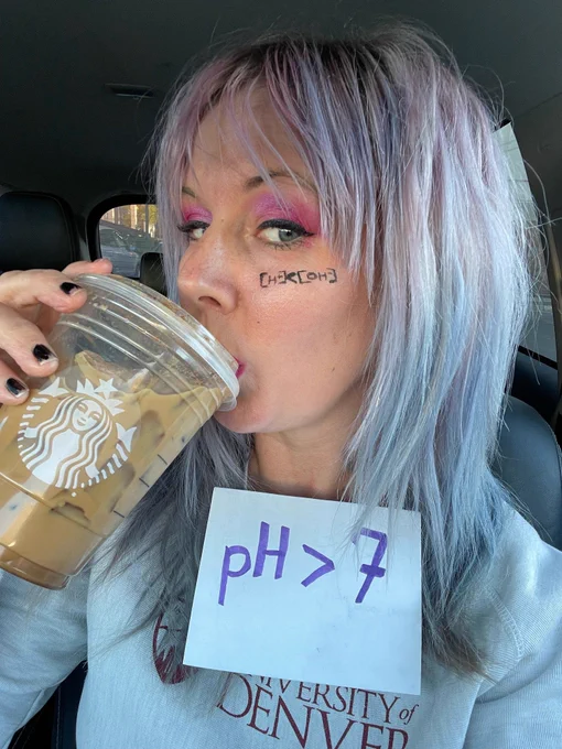A photo of a white woman with light purple almost white hair drinking a pumpkin spice latte with pink eyeshadow. On her cheek there is a formula indicating that the hydroxide concentration is greater than the proton concentration. On her chest there is a sign that says "pH>7", which indicates a basic solution.