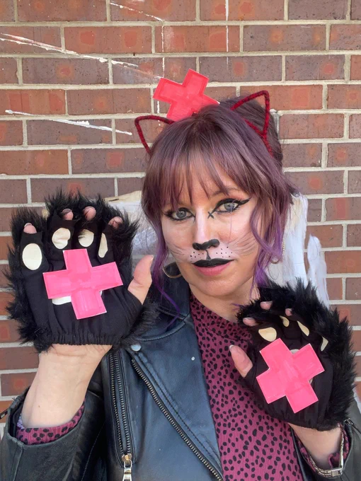 A picture of a white woman standing in front of a brick wall, dressed as a cat with red cat ears and black paws. On her paws and head there are plus signs indicating that she is a positively charged cat or a cation.