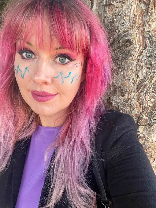 A white woman with pink hair standing in front of a tree. On her face there is a three-line nitrogen EPR signal and "I=1", which is the nuclear spin of nitrogen which results in the three hyperfine lines. 
