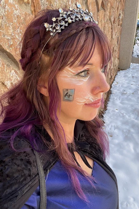 A white woman with purple hair with a crown on her head sands in front of snow lined stone wall. The symbol for the noble gas Argon is drawn on her cheek. 