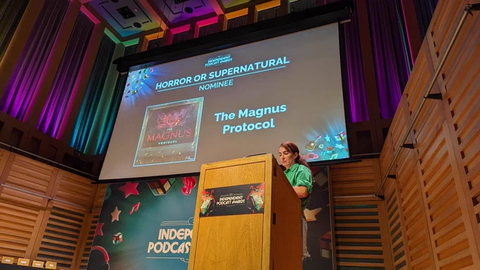 A photograph from the International Podcast Awards. A person stands at a podium on a stage, in front of a large banner for the awards and a screen showing "Horror or Supernatural Nominee: The Magnus Protocol".