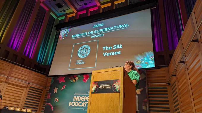 A photograph from the International Podcast Awards. A person stands at a podium on a stage, in front of a large banner for the awards and a screen showing "Horror or Supernatural Winner: The Silt Verses".