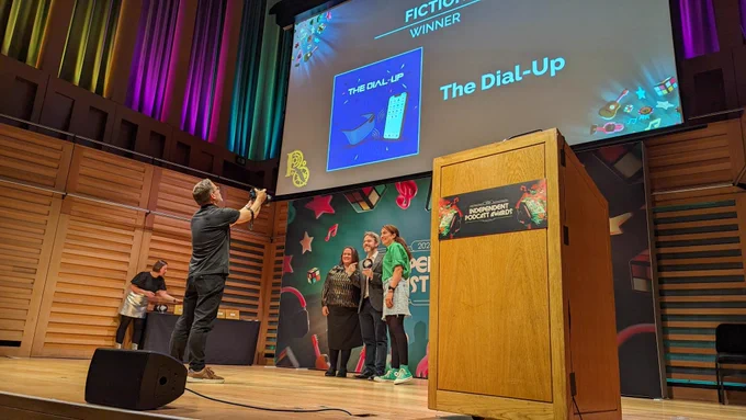 A photograph from the International Podcast Awards. A group of people stand on a stage, in front of a large banner for the awards and a screen showing "Fiction Winner: The Dial-up". They are smiling and having their photograph taken. 