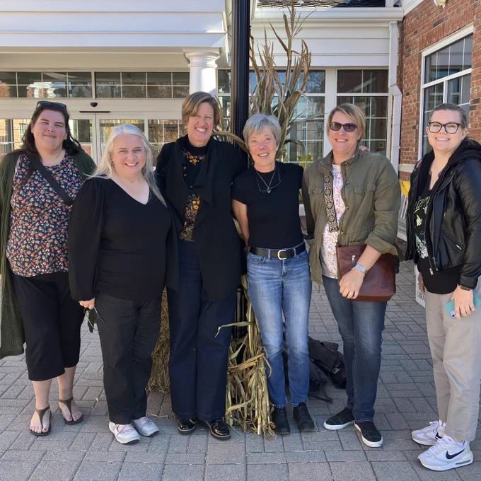 Six smiling women stand in front of a bale of hay.