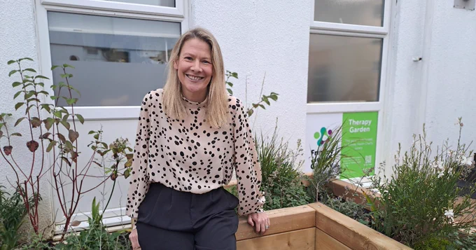 Jennie Gow smiling, sitting in front of a therapy garden sign and some green plants, wearing a polka dot shirt.