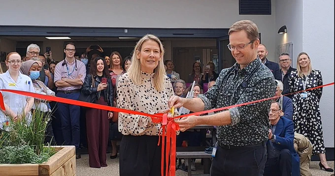Two people cutting a red ribbon at an opening ceremony, surrounded by onlookers.