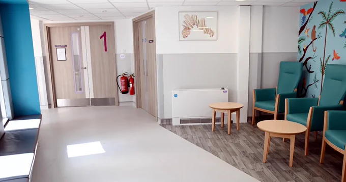 Interior view of a brightly lit hospital waiting room with numbered doors, a series of chairs aligned against a wall, small round tables, and decorative wall art featuring tropical birds.