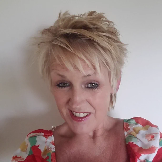 A close-up selfie of Carole Phillips, a woman with short, spiky blonde hair styled in a pixie cut. She is smiling warmly at the camera and wearing a floral top with red, green, and white colours with puffed sleeves. The background is plain and light-coloured.
