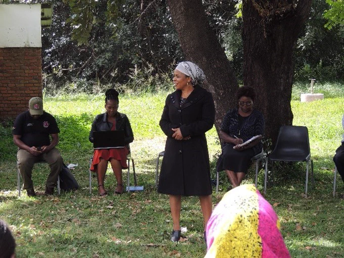 A woman standing in the middle of a circle, which is made up of people sitting on chairs. Behind her are three people sitting and in front of her is one person with the back of her head to the camera.