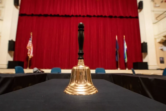 A big, shiny gold bell stands on the centre of a table in an indoor venue. The table is long and rectangular, covered in a black tablecloth and there are chairs placed around it. Behind them is a stage is hung with tall, heavy, red curtains.