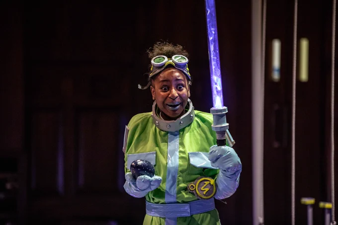 A woman dressed in a yellow boiler suit holds a plastic light sabre up in front of her. She is wearing goggles on her head and has an egg in the other hand. 