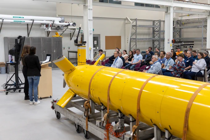 Two people present at a podium in front of a seated audience. In the foreground, a large yellow autonomous underwater vehicle is strapped down to a wheeled dolly.