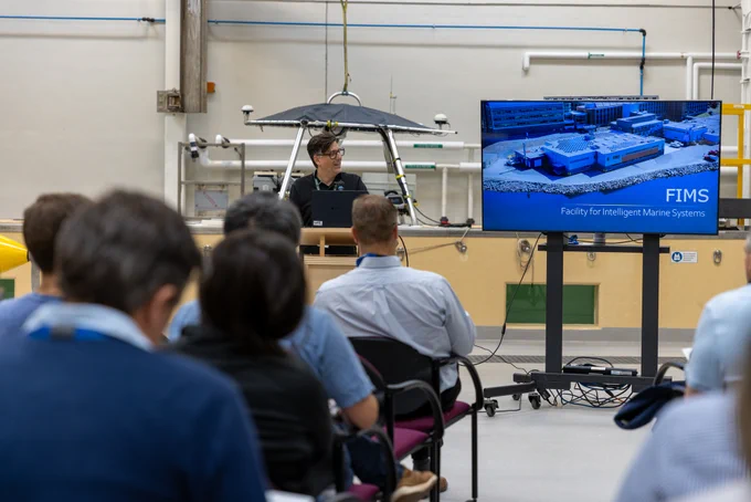 A presenter at a podium addresses a seated audience. There is an image of a building on a screen with the words “FIMS: Facility for Intelligent Marine Systems.” In the background, a metal frame with scientific equipment mounted on it is hanging from a strap from the ceiling.