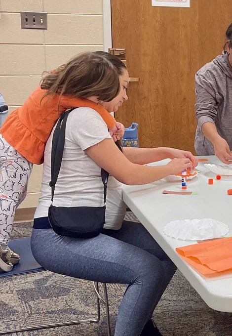A child leans over an adult's shoulders as the adult glues tissue paper to a coffee filter