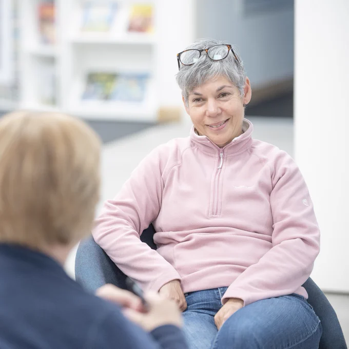 A woman in a pink zip up fleece sat in a blue chair, speaking to someone in front of her.