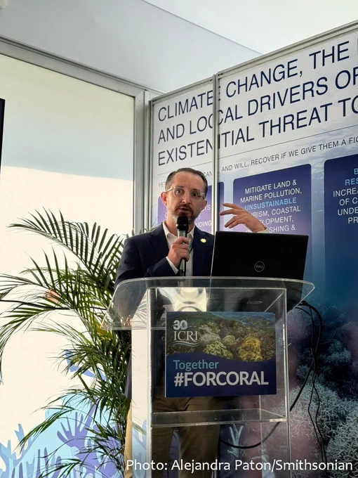 A man wearing a suit and glasses holds a microphone and speaks behind a lectern. The front of the lectern is decorated with a photo of coral and the words "Together #ForCoral". Credit: Alejandra Paton/Smithsonian