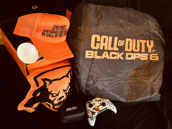 An orange trucker hate, orange pillow, small black case, Black ops Xbox controller, and a black bomber jacket on a black background.