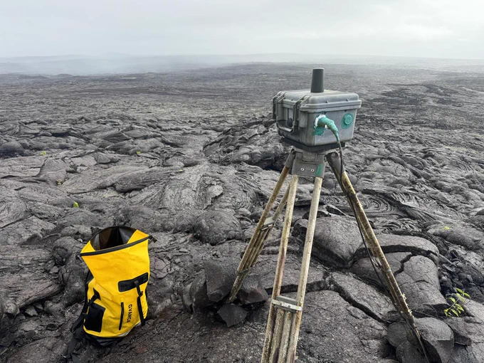 A webcam sits atop a tripod on a bed of black lava. Instruments to fix the camera are nearby in a yellow waterproof bag. The R3cam is a webcam on the east flank of Pu‘u‘ō‘ō, and it monitors areas downrift of Pu‘u‘ō‘ō. USGS geologists visited the site on October 23, 2024 to dry out the camera enclosure and add new desiccant. USGS photo by M. Patrick.