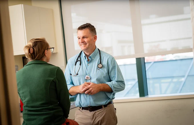 A healthcare professional wearing a stethoscope converses with a patient in a medical office, both smiling.