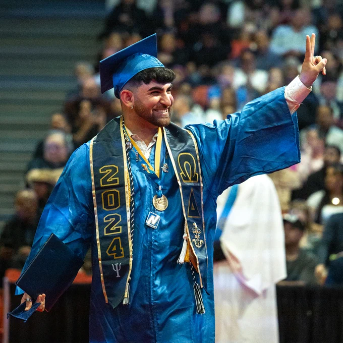 A person wearing commencement regalia looks to the left and gestures with arm in the air