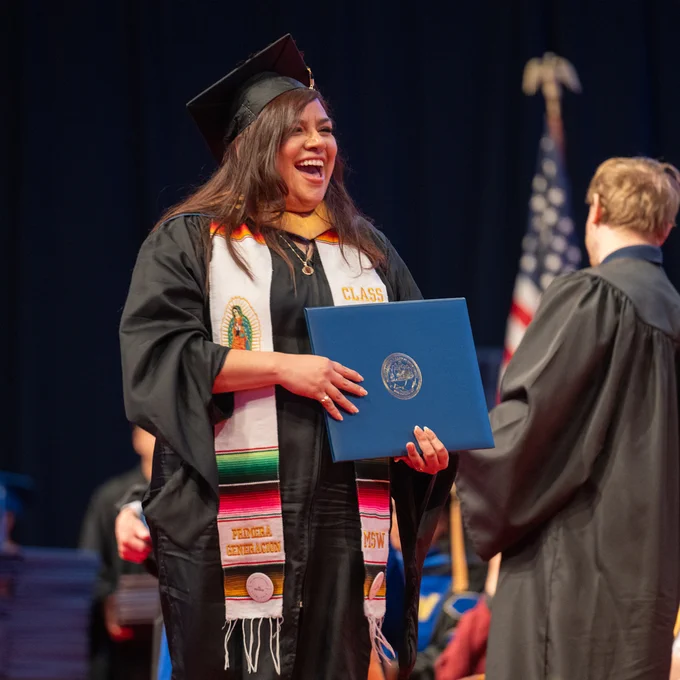 A person in commencement regalia with a big smile crosses the stage after receiving their diploma.