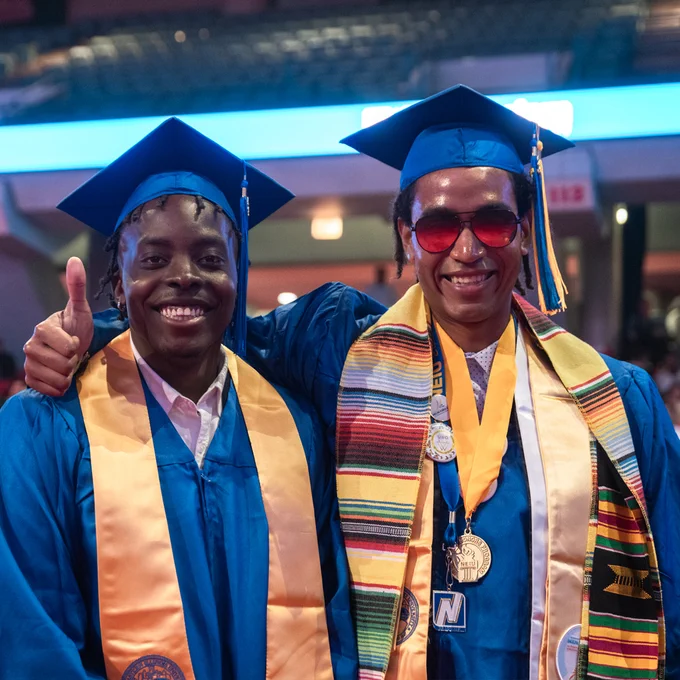 Two people wearing commencement regalia stand close together while smiling at the camera.