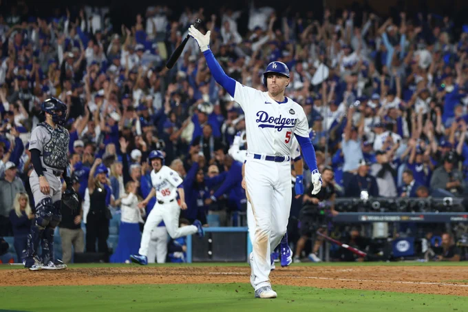 Freddie Freeman tosses his bat after his World Series Game 1 walk-off grand slam. He wears a home white Dodgers uniform with blue and red lettering. The Dodger Stadium crowd goes nuts in the background.