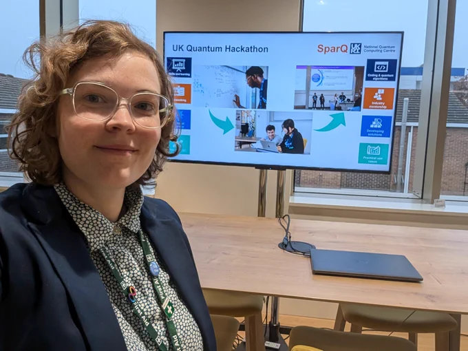 A white femme(ish) presenting person wearing glasses and a navy blazer smiling in front of a large monitor about the UK quantum Hackathon.