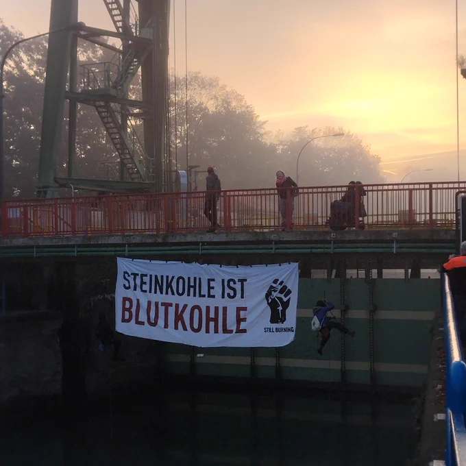 Ein Transparent hängt an einer Brücke direkt über der Schleusenzufahrt. Darauf steht: "Steinkohle ist Blutkohle." Auf der Brücke stehen mehrere Aktivist*innen.