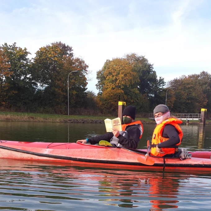 Zwei rote Kayaks mit Personen in orangenen Warnwesten darin. Eine Person liest in einem Buch.