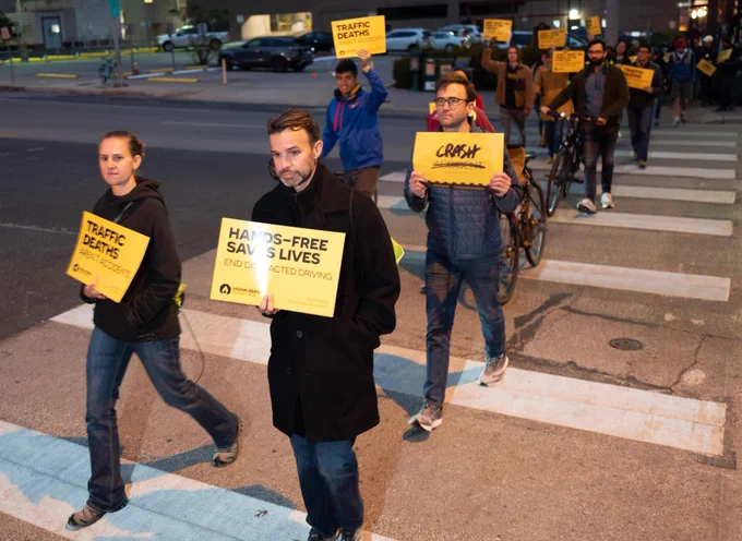 A group of people walking across a crosswalk holding yellow signs that say “traffic deaths are not accidents” and “Hands-free saves lives”