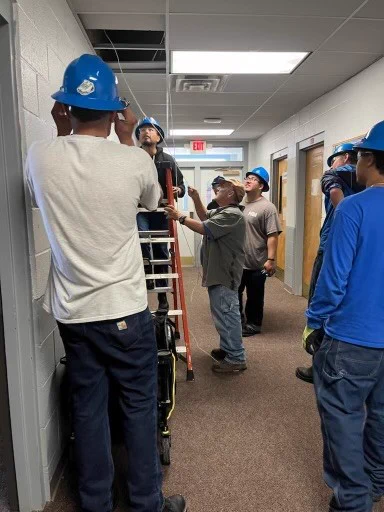 A group of Electrical students gather around their instructor while looking at electrical cords in the ceiling.