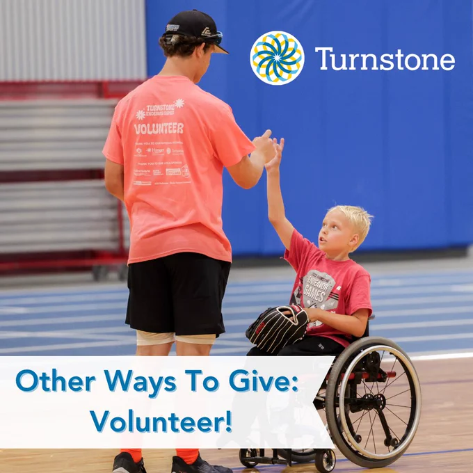 Teen boy dressed in a pink shirt that says volunteer on the back gives a high five to a young boy in a wheelchair wearing a baseball mitt. 