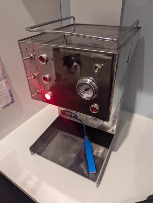 A coffee machine with some kind of micro computer plugged into it. It has several lights on the front of it and one is lit up red.