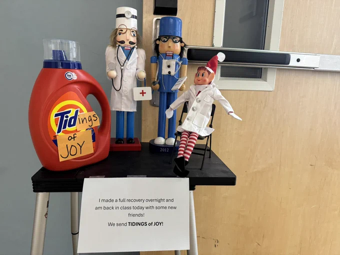 Two nutcrackers and an elf dolls dressed up as doctors and nurses on the table next to Tide laundry detergent. A message on a sheet of paper says: I made a full recovery overnight and am back in class today with some new friends. We send TIDINGS of JOY!