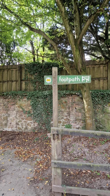 The image shows a footpath sign mounted on a wooden post. The sign has a green background with the word "footpath" written in white, accompanied by an arrow pointing to the right. Above the word "footpath" is a circular emblem of two opposing yellow arrows indicating that this is part of the route of the Lambourn valley way. The post is set against a rustic backdrop of a stone wall overgrown with ivy, with additional trees and greenery surrounding the area, giving it a natural and inviting look. Fallen leaves and soil cover the ground near the base of the post.

