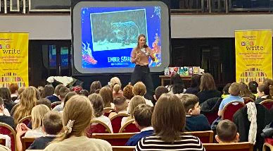 
Shot of audience in theatre with presenter on stage. Large screen and bright yellow displays saying Wee Write are on the stage
