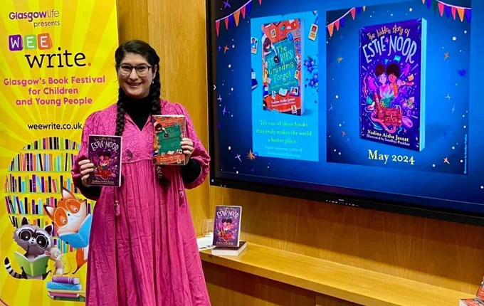 Author Nadine Aisha Jassat holding up two of her books in front of large screen, also showing books, and yellow Wee Write display stand