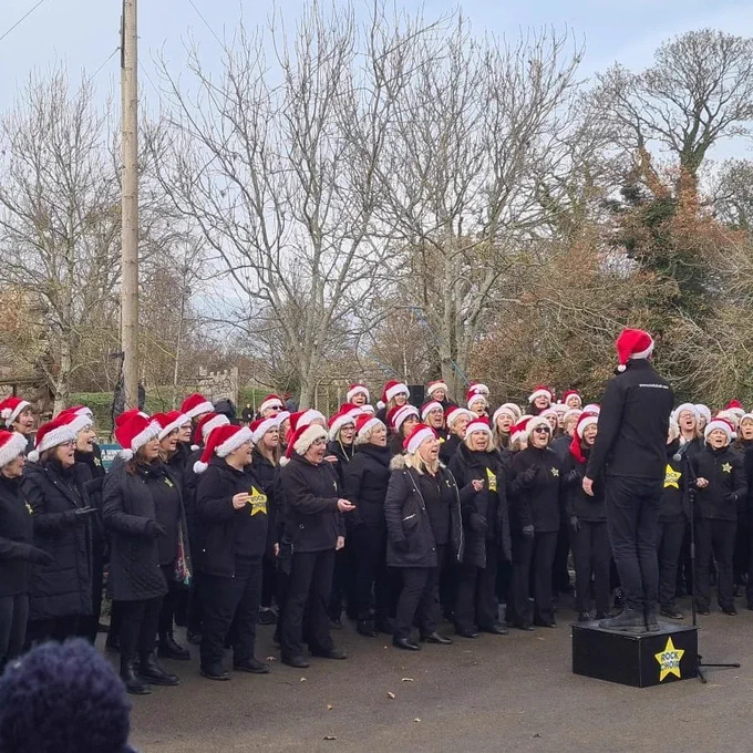 A choir wearing Santa hats and black outfits performs outdoors, conducted by a person standing on a small black platform.