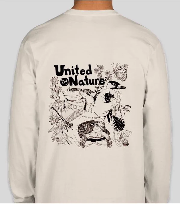 A crème colored shirt showing hands holding fabulous critters (bullfrog, box turtle, and red-cockaded woodpecker), a dragonfly and butterfly along with plants and flowers, and sports our 2025 theme “United by Nature