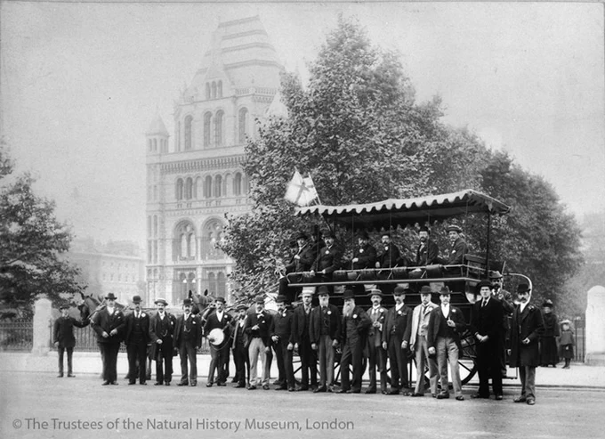 A horse-drawn bus sits on the road outside the museum building. Seven men can be seen sitting in the vehicle and eighteen stand outside.
