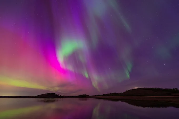 An aurora, glowing magenta and green in the sky, shines over a large lake with small islands and hills in the background. Credit: Gunjan Sinha