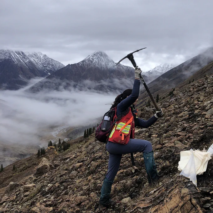 Earth scientist Ayesha Landon-Browne wields a geological hammer over a rocky hillside. 