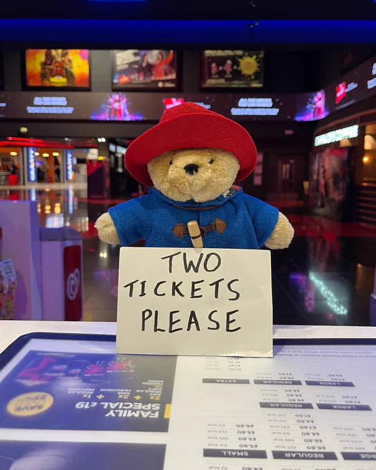 Paddington Bear plush toy standing at a cinema counter, wearing a red hat and blue coat, with a sign reading 'TWO TICKETS PLEASE.' Movie posters are visible in the background.