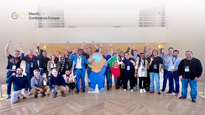 A diverse group of attendees stands together in a spacious venue, smiling and raising their hands in celebration. In the center, there is a large mascot figure wearing an astronaut suit. The background features modern architecture with wooden flooring.